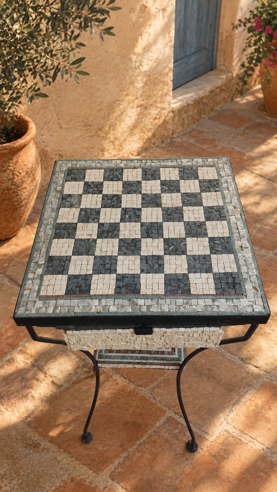 A Knight's Table mosaic chess table outdoors, full tabletop and iron-leg base with the decorative shelf board visible beneath